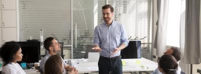 A man in a light blue button-down shirt and glasses stands at the center of a modern office, gesturing while speaking to five colleagues seated around him in an informal team discussion.
