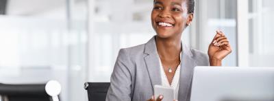 smiling businesswoman in a gray blazer sits at her desk holding a smartphone, with a laptop open in front of her in a bright office setting.