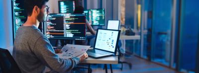 A software developer sits at a desk reviewing a printed UI wireframe while working across multiple monitors displaying code in a dimly lit office.