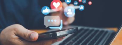 A person holds a smartphone above a laptop while social media icons — including likes, hearts, messages, and profile icons — float above the screen.