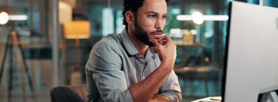 A young professional man with glasses sits at a desk in a modern office at night, looking thoughtfully at a computer monitor