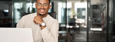 A young professional man wearing glasses sits at a desk with a laptop and open notebook, smiling confidently in a modern office environment.
