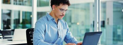 A young man with curly hair and glasses sits at a desk in a modern office, typing on a laptop.
