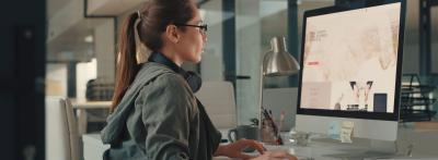 A woman with headphones around her neck sits at a desktop computer in a modern office, browsing a website on a large monitor.