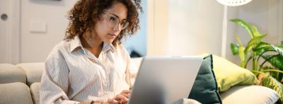 A young woman with curly hair and glasses works on a laptop 