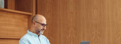 A middle-aged man with glasses and a short beard sits at a desk working on a laptop in a warmly lit office with wood-paneled walls.