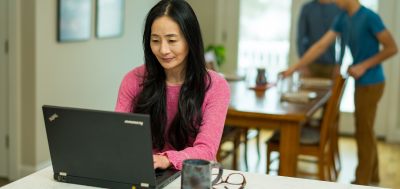 Asian woman at laptop sons in background setting dinner table