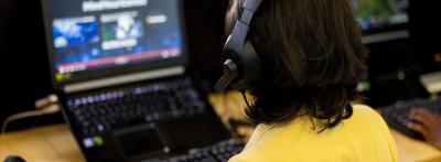A person wearing a headset works at a laptop displaying video game design software, with additional monitors visible in the background.