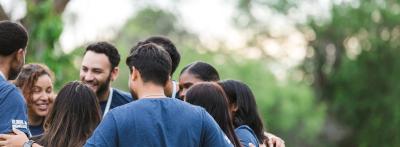 A diverse group of colleagues in matching t-shirts stand in a huddle outdoors with arms around each other, representing community and social connection.