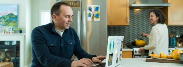Man at laptop in kitchen with wife at stove