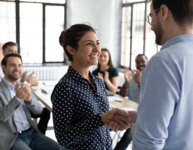 Standing woman shaking male manager's hand in front of colleagues sitting at table