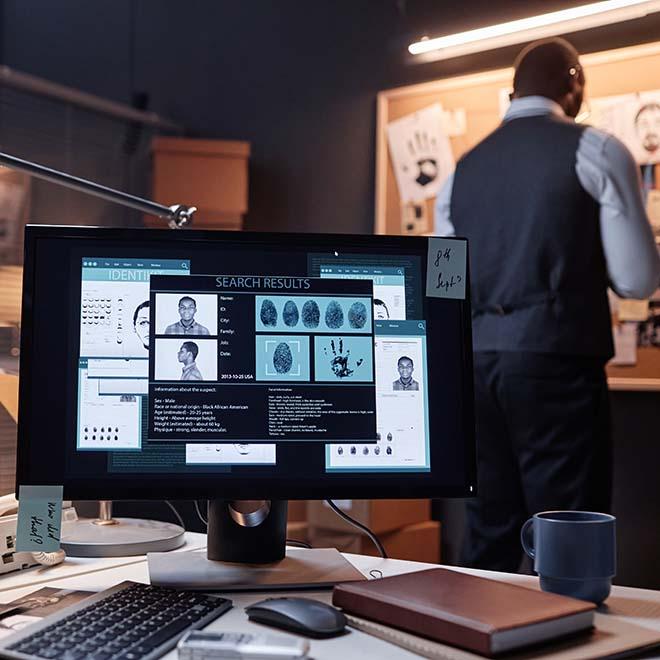 Detective in a room looking at a bulletin board with a suspect on a computer monitor